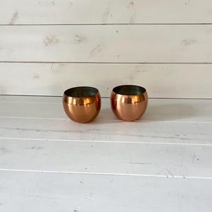 May include: Two small, spherical cups with a polished copper exterior finish and a dark, matte interior. The cups have a rounded, bowl-like shape with a wide opening at the top and a smooth, reflective metallic surface throughout.