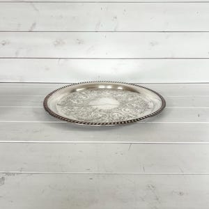 May include: A silver-colored, round serving tray with an ornate floral design in the center. The tray has a beaded edge and is set against a white wooden background. The tray is likely made of metal and is suitable for serving food or displaying items.