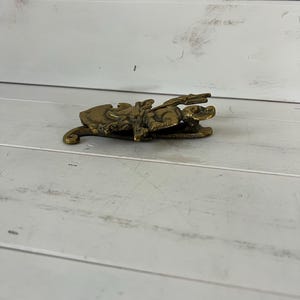 Może przedstawiać: Szczeg&oacute;łowa, antyczna mosiężna figurka mitycznego stworzenia, być może smoka lub gryfa, z misternymi detalami. Figurka leży na białej drewnianej powierzchni. Mosiądz ma ciepłą, postarzaną patynę.