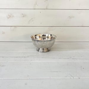 May include: A polished silver-toned metal bowl with a smooth, rounded basin and a tiered pedestal base. The bowl features a reflective surface and a simple, clean design with a wide opening and a sturdy, circular footed bottom for stability.