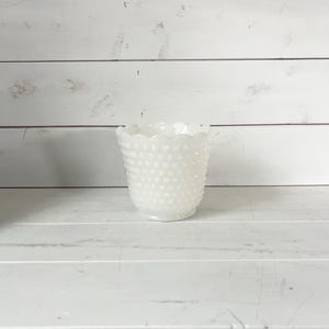 May include: A white hobnail glass vase with a scalloped rim. The vase has a textured surface with small, rounded bumps. The background is a white wooden surface.
