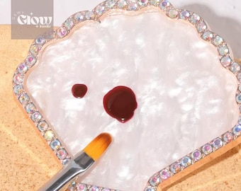 Puede incluir: Una paleta blanca en forma de concha con un borde dorado y diamantes de imitación alrededor del borde. Hay dos pequeñas gotas de pintura roja en la paleta, y un pincel descansa en el borde.