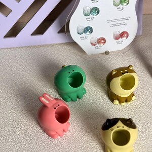 May include: Four colorful animal-shaped fingertip ring holders. The holders are a green frog, a pink bunny, a brown dog, and a yellow cat. Each holder has a large open mouth to hold rings.