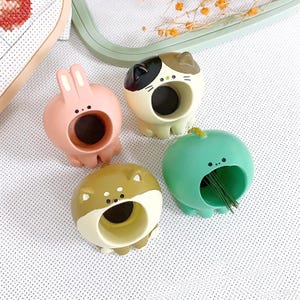 May include: Four ceramic needle minders shaped like animals. A pink bunny, a brown and white cat, a green dinosaur, and a brown dog.
