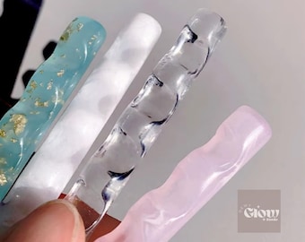 May include: Close-up of four nail art tools in various colors. The first is a light blue with gold flecks, followed by a white, a clear with dark blue swirls, and a pink. The tools are held by a person's finger.