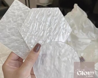May include: A collection of white, pearlescent coasters in various geometric shapes: a diamond, a hexagon, and a circle. The coasters have a wavy, textured surface. The image also includes the logo for "Dewi Glow + Studio".