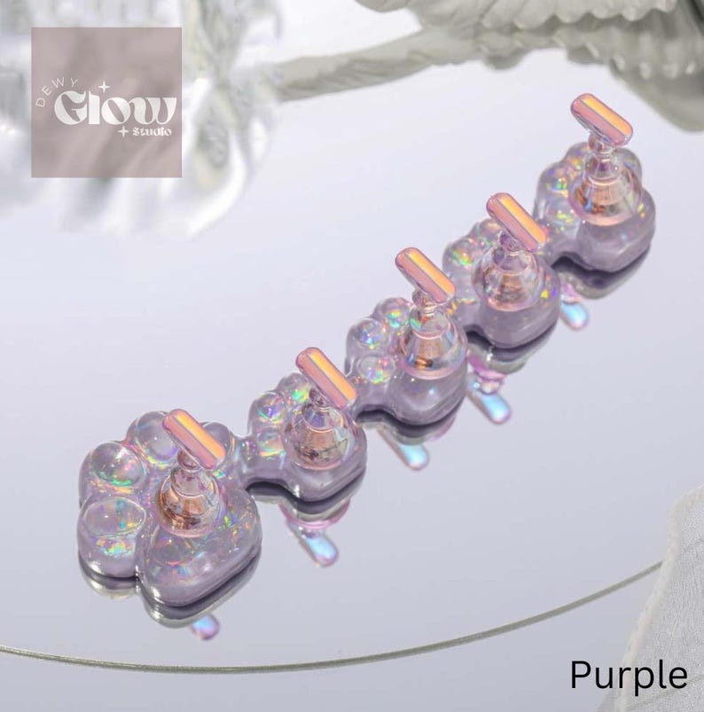 Puede incluir: Un conjunto de cinco soportes para esmalte de u&ntilde;as de color morado iridiscente con un dise&ntilde;o de huella de pata. Cada soporte tiene una peque&ntilde;a parte superior redonda transparente con un soporte para esmalte de u&ntilde;as de metal. Los soportes est&aacute;n dispuestos en una fila sobre una superficie reflectante. El texto "DEWY GLOW STUDIO" es visible en el fondo.