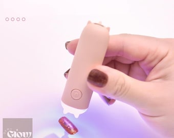May include: A pink handheld UV lamp with a cat-shaped design. The lamp has a white button on the side and a white light at the top. The lamp is being held by a hand with dark brown nail polish.