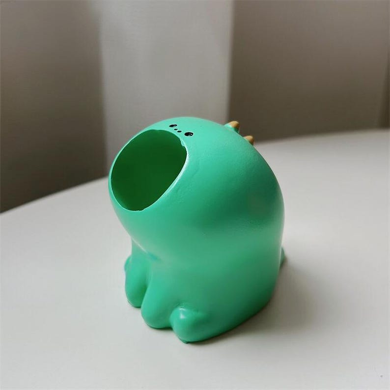 May include: A green ceramic planter shaped like a cartoon monster with a large open mouth. The planter has two small black eyes and a light brown nose.
