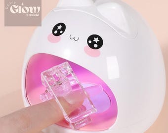 May include: White cat-shaped UV nail lamp with a pink interior and a clear plastic finger holder. The lamp has a cute cartoon face with black eyes and pink cheeks.