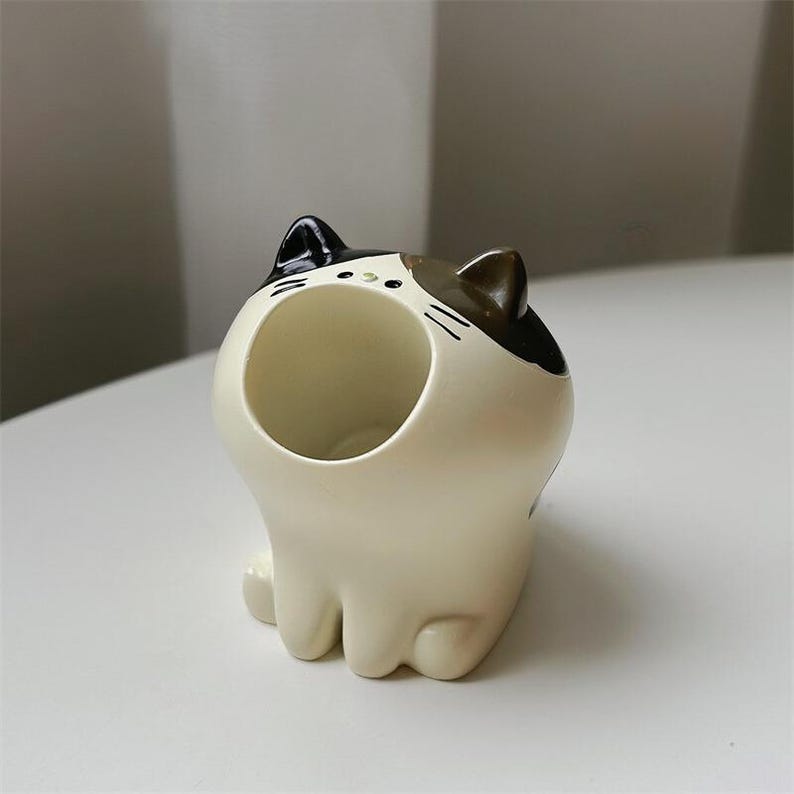 May include: A white ceramic cat figurine with black and brown markings. The cat has a large open mouth, forming a small container.