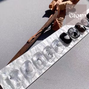 Pode incluir: Um conjunto de unhas posti&ccedil;as transparentes com desenhos de glitter prateado, incluindo borboletas, estrelas e um dinossauro. Algumas unhas s&atilde;o pretas com detalhes de cora&ccedil;&atilde;o prateado. As unhas s&atilde;o exibidas em uma superf&iacute;cie marmorizada branca e cinza.