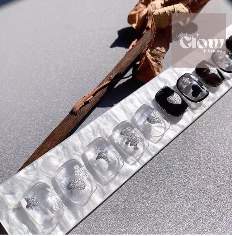 Pode incluir: Um conjunto de unhas posti&ccedil;as transparentes com desenhos de glitter prateado, incluindo borboletas, estrelas e um dinossauro. Algumas unhas s&atilde;o pretas com detalhes de cora&ccedil;&atilde;o prateado. As unhas s&atilde;o exibidas em uma superf&iacute;cie marmorizada branca e cinza.