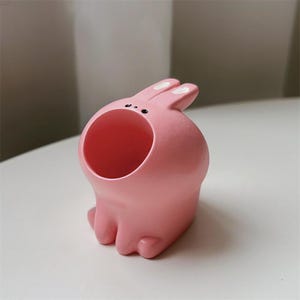 May include: A pink ceramic bunny planter with a large opening for planting. The bunny has small black eyes and white ears.