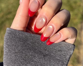 May include: Close-up of a hand with oval-shaped nails painted with a nude polish and a red half-moon design at the base of each nail.