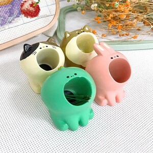 May include: Four colorful ceramic needle minders shaped like animals. A green frog, a pink bunny, a brown and white cat, and a brown dog. Each minder has a hole in the top for holding needles.