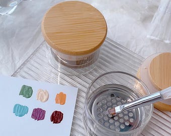 May include: Two clear glass jars with bamboo lids and a metal mesh insert. One jar has a paintbrush resting on the mesh. A white card with six small paint swatches in different colors is in the background.