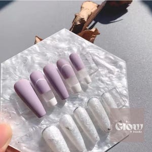 Pode incluir: Um conjunto de unhas posti&ccedil;as foscas lavanda com um detalhe branco e um design de linha branca. Abaixo, unhas brancas com um design marmorizado. As unhas s&atilde;o exibidas em uma superf&iacute;cie branca texturizada.