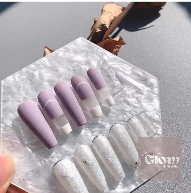 Pode incluir: Um conjunto de unhas posti&ccedil;as foscas lavanda com um detalhe branco e um design de linha branca. Abaixo, unhas brancas com um design marmorizado. As unhas s&atilde;o exibidas em uma superf&iacute;cie branca texturizada.