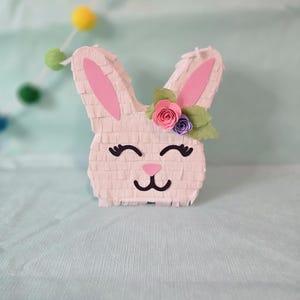 May include: A white bunny piñata with pink ears and a flower crown. The crown has pink, purple, and green flowers.