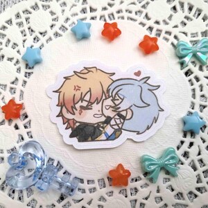May include: A sticker featuring two anime-style characters embracing, with one character's face showing an angry expression. The sticker is surrounded by blue and orange star-shaped beads, and teal bow-shaped beads. A clear, blue, heart-shaped charm is also present.