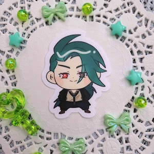 May include: A sticker featuring a cartoon character with teal hair and red eyes, dressed in a black outfit. The sticker is surrounded by green embellishments, including bows, stars, and beads, on a white lace doily.
