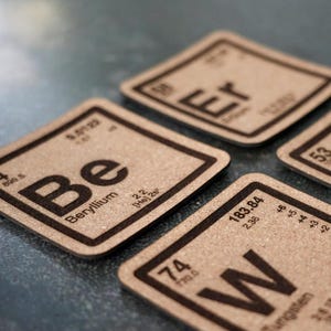 May include: A collection of square coasters with a science theme. Each coaster features a different element from the periodic table, with the element symbol, atomic number, and name printed in a dark brown color. The coasters are made of a light brown material.