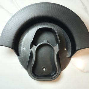 May include: A black 3D printed wall mount with a curved design and a circular base. The mount has a cutout for a device, such as a phone or a tablet.