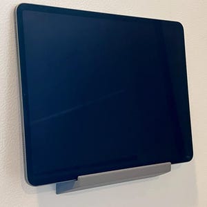 May include: A dark screen tablet is mounted on a gray holder against a white wall. The tablet is rectangular with rounded corners and a thin silver frame. The holder is a simple, angled design.
