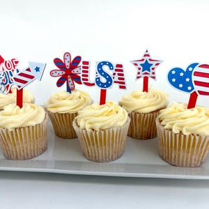 May include: Six cupcakes with yellow frosting on a white tray, each topped with a patriotic decoration. The toppers feature fireworks, a star, a flower, the letters "USA", and heart shapes, in red, white, and blue. The cupcakes are ideal for a celebration.