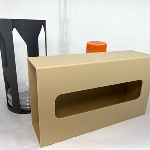 May include: A beige rectangular box with a long, oval opening, next to a black holder with a circular opening. An orange cap sits on top of the box, and a clear cylinder is in the background. The items appear to be laboratory equipment.
