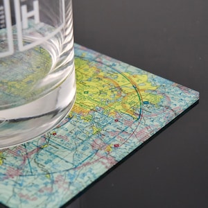 May include: A square coaster featuring a detailed aeronautical sectional chart of the Atlanta region with yellow, blue, and red navigational markings. A clear glass with white printed text sits partially on the coaster surface.