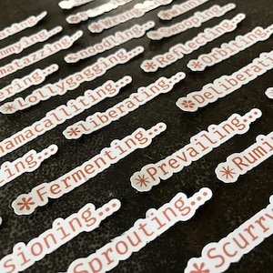 May include: A collection of white stickers with red text, featuring various words like "Nucleating," "Shimmying," and "Razzmatazzing." Each sticker has a small asterisk design. The stickers are arranged on a dark surface.