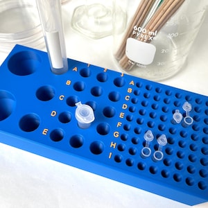 Puede incluir: Estante de plástico azul con filas de agujeros redondos para sostener pequeños tubos. El estante está etiquetado con las letras A a I y los números 1 a 12. Hay varios tubos de plástico transparente con tapas en los agujeros.
