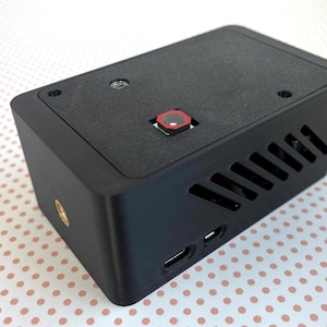 May include: A black rectangular electronic device with a red button and ventilation slots. The device has a textured surface and a small port on the side. A gold-colored screw is visible on the side.