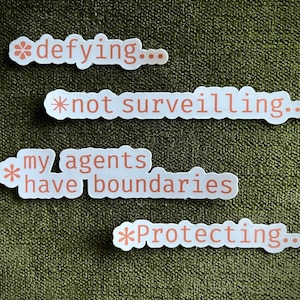 May include: Four white stickers with text in a light orange color. The stickers say "defying...", "not surveilling...", "my agents have boundaries", and "Protecting...". The stickers are on a textured, olive green surface.