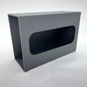Puede incluir: Una caja rectangular gris con una gran abertura rectangular redondeada. La caja está hecha de un material sólido y tiene un acabado mate. La abertura es oscura, creando un contraste con el exterior gris.