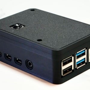 May include: A black plastic case for a Raspberry Pi computer with a camera lens on the top and multiple ports on the side.