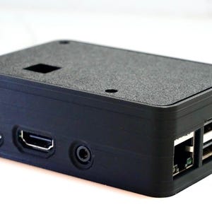 May include: A black plastic case with multiple ports, including USB, HDMI, and Ethernet. The case has a textured surface and is designed to protect a small electronic device.