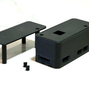 Peut inclure: Un boîtier en plastique noir avec une ouverture rectangulaire et un support en plastique noir séparé avec quatre pieds. Le boîtier a une série de petits trous sur le côté.