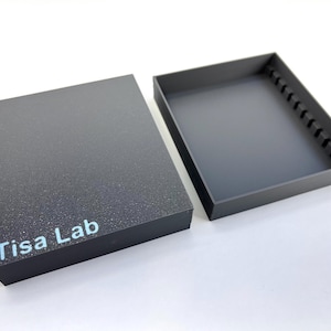 Puede incluir: Una caja rectangular negra con tapa a juego, abierta para revelar una bandeja interior. La caja tiene una superficie texturizada y las palabras "Tisa Lab" están impresas en azul claro. La bandeja tiene una serie de pequeños divisores negros.