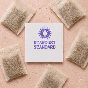 May include: Five rectangular tea bags with visible dark tea leaves arranged around a central square white card. The card features a purple sun-like icon above the text STARDUST STANDARD in a bold, sans-serif font, centered on the white surface.
