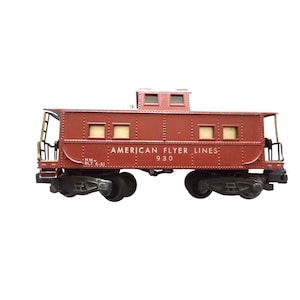 May include: A maroon model train caboose with the text "AMERICAN FLYER LINES: 930" printed on the side. The model has black wheels and a small observation cupola on top. The caboose has small windows and metal railings.