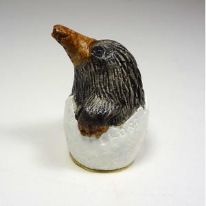 以下が含まれることがあります： ひびの入った卵から出てくる鳥の陶器の彫刻。鳥は茶色のくちばし、黒い羽、白い卵殻の台座を持っています。白い背景にあります。
