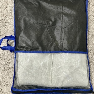 Op de afbeelding: Een rechthoekige opbergtas met een blauwe stoffen rand en handvat. Het bovenste gedeelte is gemaakt van zwart vliesmateriaal, terwijl het onderste gedeelte een doorzichtig plastic venster heeft dat een gevouwen wit textielstuk in de tas toont.