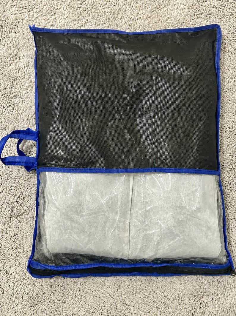 Op de afbeelding: Een rechthoekige opbergtas met een blauwe stoffen rand en handvat. Het bovenste gedeelte is gemaakt van zwart vliesmateriaal, terwijl het onderste gedeelte een doorzichtig plastic venster heeft dat een gevouwen wit textielstuk in de tas toont.