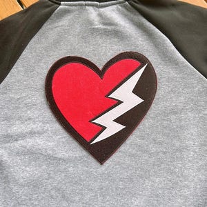 Puede incluir: Una chaqueta de béisbol gris y negra con un corazón rojo y blanco con un diseño de rayo en la espalda.