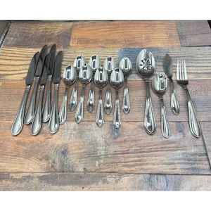 May include: A collection of stainless steel silverware, including knives, spoons, and forks, arranged on a wooden surface. The silverware has a simple, elegant design with rounded handles. The set includes serving spoons and a butter knife.