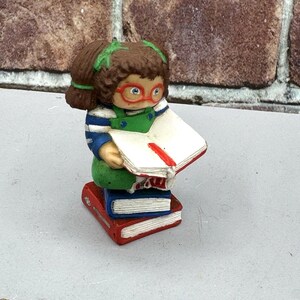 Peut inclure: Une petite figurine représentant un personnage aux cheveux bruns avec des couettes, portant des lunettes rouges, un haut rayé bleu et blanc et une salopette verte. Le personnage est assis sur deux livres empilés et tient un livre ouvert.