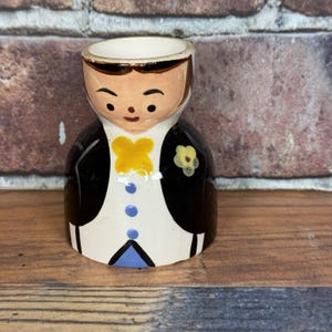 May include: A vintage ceramic egg cup shaped like a man in a tuxedo. The egg cup has a white shirt, black jacket, and a yellow bow tie. The face is painted with brown hair and a smiling expression.